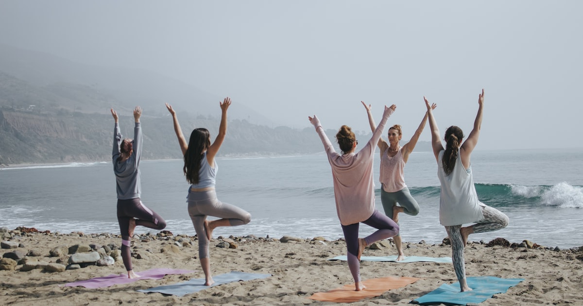 Group of women in their 30s and 40s practicing tree pose yoga on a beach, representing wellness during early perimenopause